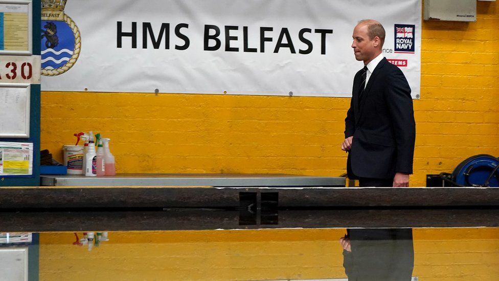 Prince William standing in front of a sign reading HMS Belfast