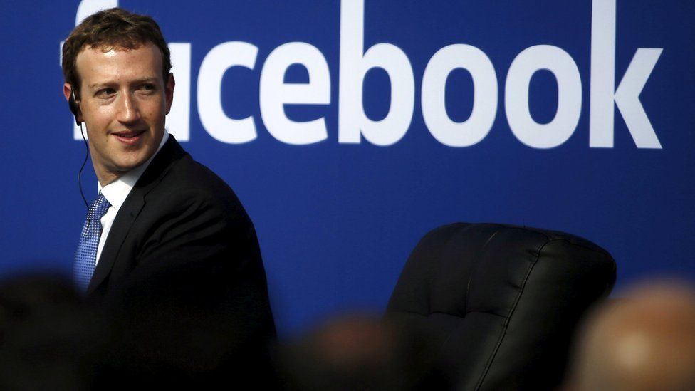 Mark Zuckerberg, wearing a suit and audio earpiece, sits in front of a a Facebook logo