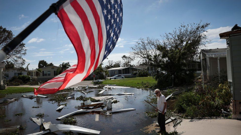 A man cleans debris from Hurricane Ian with American flag waving through.