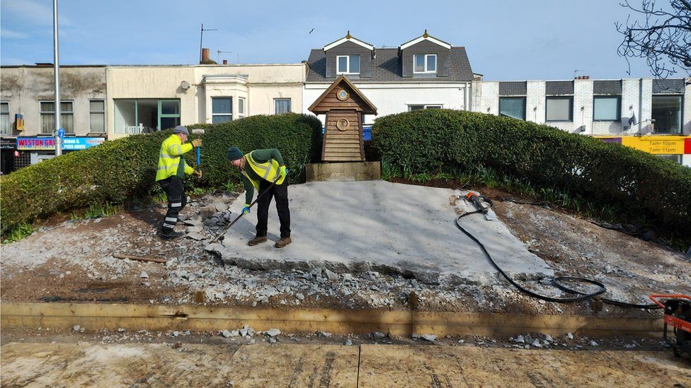 Two workmen removing the concrete covering beside the flower clock