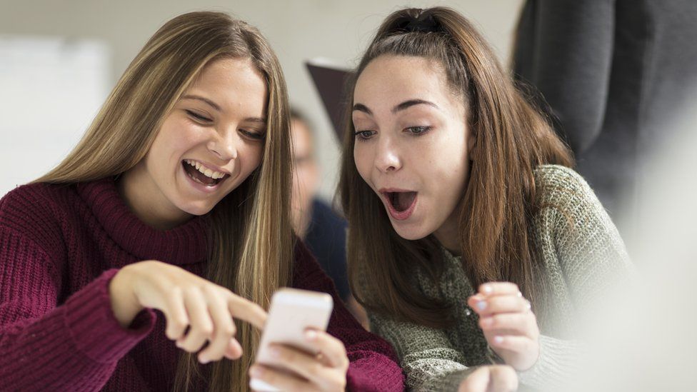Two teenage girls point and laugh at a smartphone screen in this stock photo