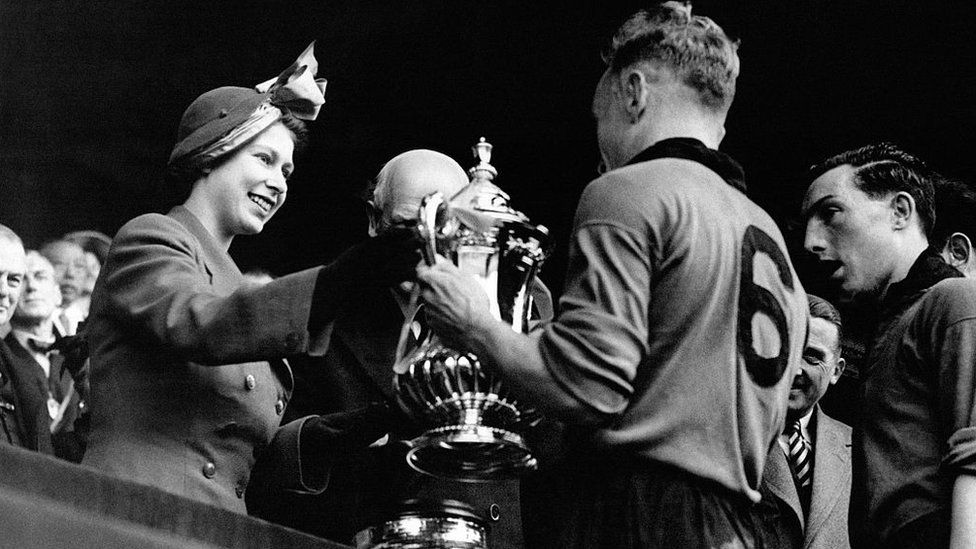 A black and white photo of smiling queen handing over the trophy