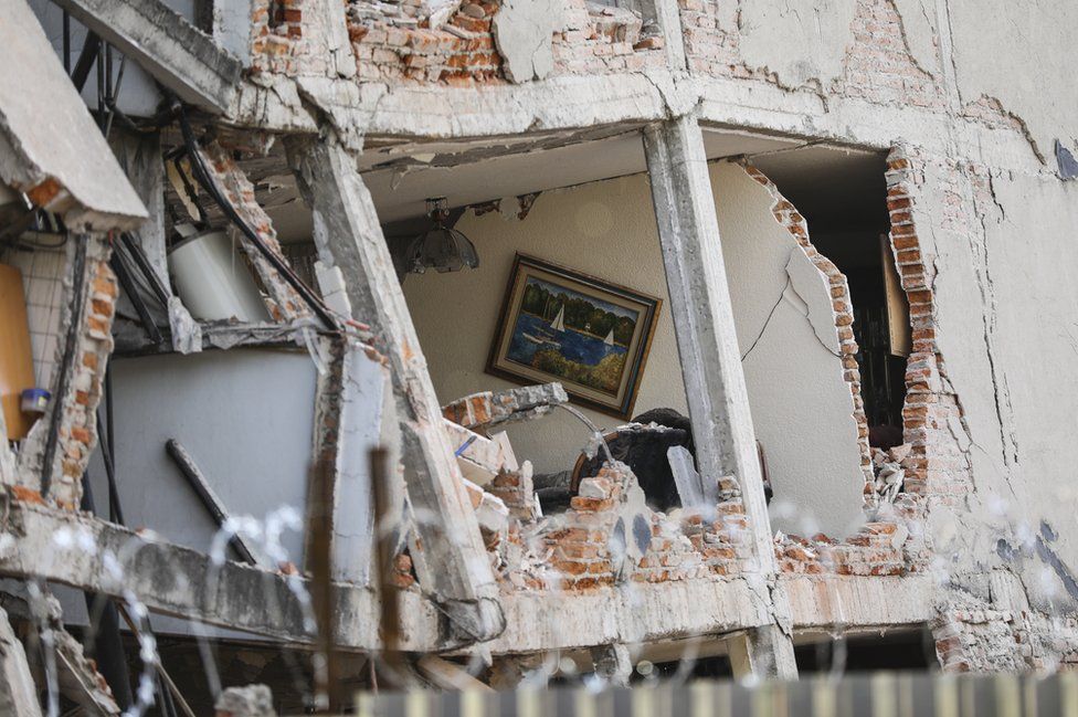 A damaged building with a hole in the exterior reveals a painting hanging on the wall inside - showing a tranquil boating scene