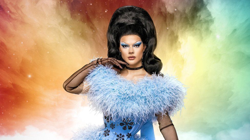 Gothy Kendoll in a promotional photo for Drag Race UK vs The World. Gothy is a 26-year-old drag queen, wearing an enormous dark wig and sky-blue corseted dress embellished with feathers, black flowers and black lace gloves. She wears blue eye shadow to match her dress and holds her gloved right hand to her chin. The background is a rainbow coloured galaxy scene