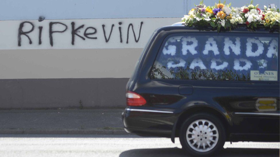 A funeral car carrying the coffin of Kevin McGuigan Sr passes graffiti on a wall that reads: 'RIP Kevin'