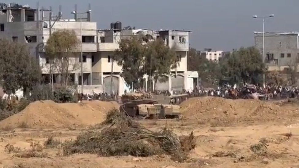 A still of a video showing people walking up Salah al-Din road with a tank nearby