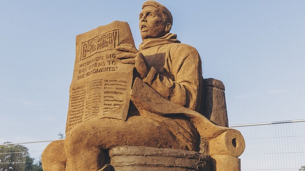 A mud and sand sculpture of a man on a toilet, reading a newspaper