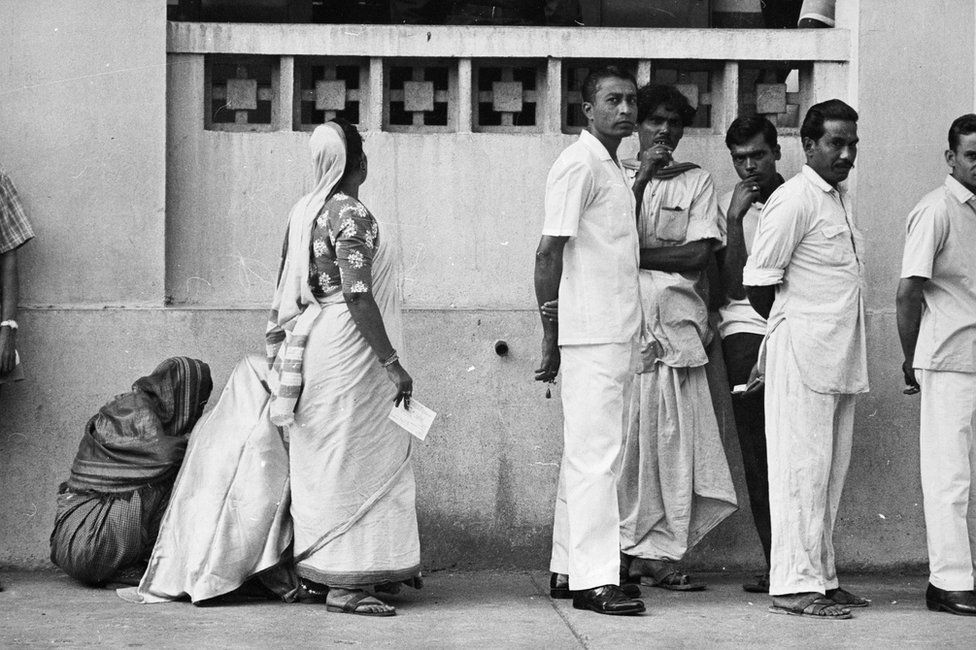 A woman in a queue to vote, turns her face to the wall as men face the camera in 1967