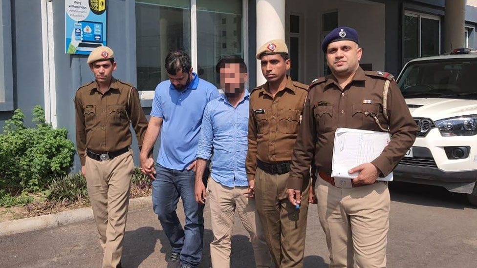 Two men in blue shirts are flanked by Indian police officers outside a building