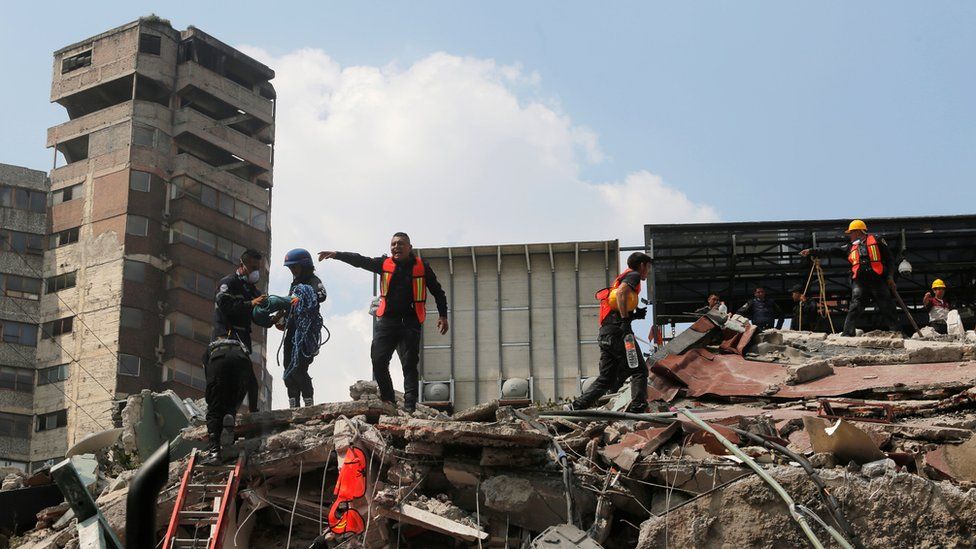rescuer in high vis jacket pointing and shouting, while standing on debris next to a tall building