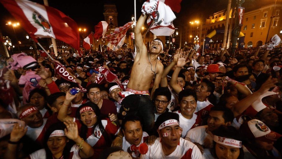 A large crowd of Peruvian fans wave their flags as a shirtless man is carried shoulder high