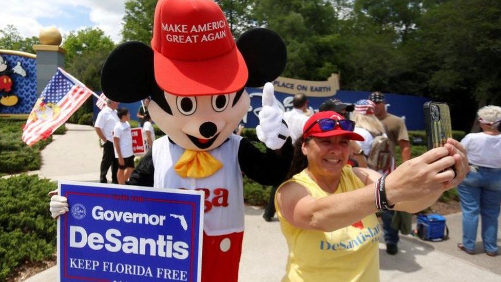 A person in a mouse costume holds a DeSantis campaign poster and takes selfies with a supporter in April 2022