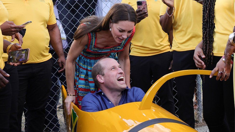 Prince William sits in a bobsleigh while his wife pushes