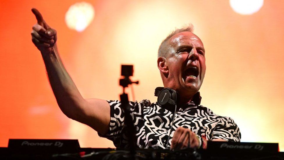 Fatboy Slim in the middle of a DJ set. He's bathed in the orange glow of a stage light, and is pointing one finger up in the air as he energetically yells encouragement to the crowd. He's got a pair of over-ear headphones looped around his neck, and a glimpse of his black-and-white, zebra patterned shirt is visible, as is the top of his DJ desk.