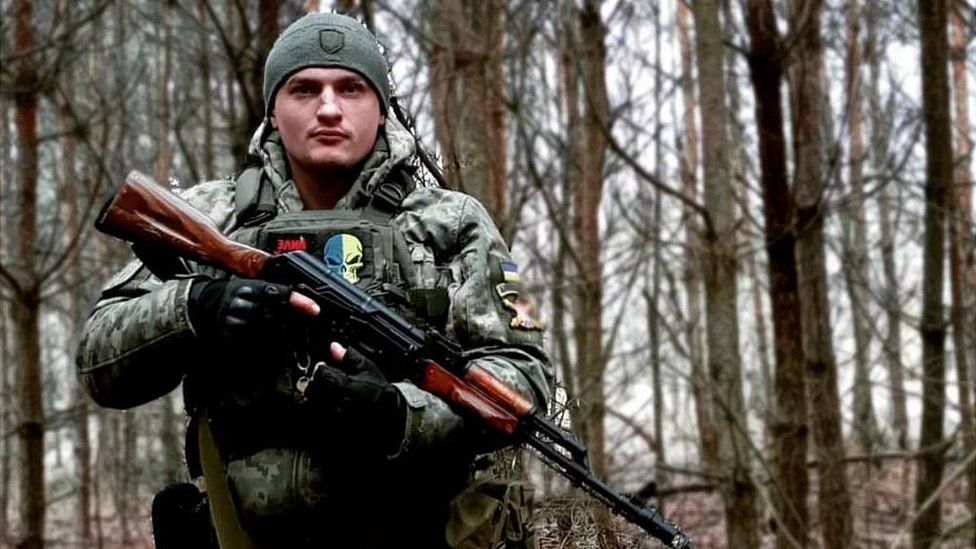 Serhiy poses for a picture in a forest, wearing combat uniform and holding assault rifle