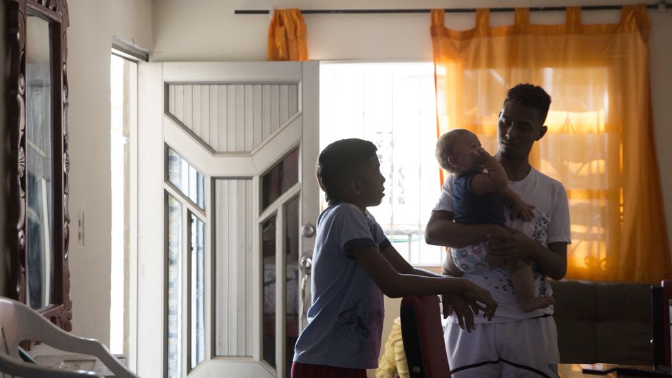 League leader Eidavis Montes' husband and son care for her baby in the living room of her house.