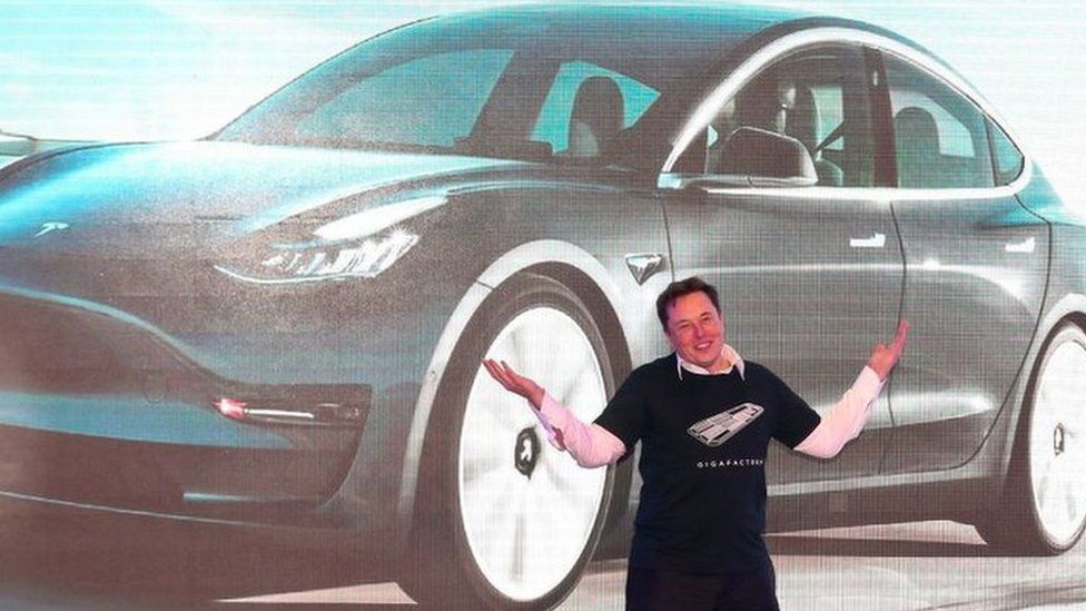 Elon Musk on stage in front of an image of a Tesla Model 3