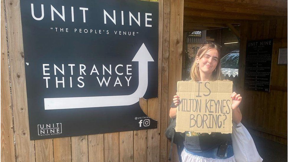 Anouska Lewis holding a sign that says "Is Milton Keynes Boring?" outside Unit Nine music venue
