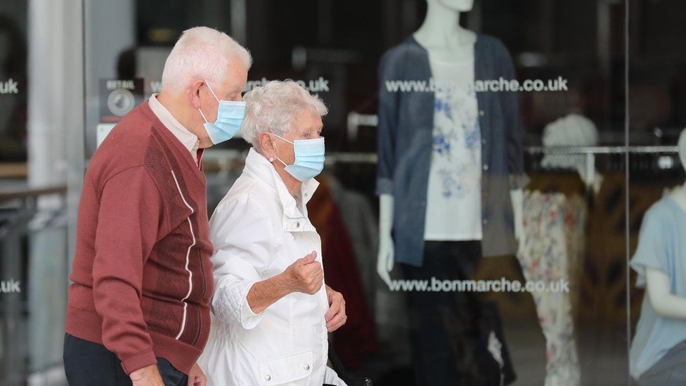 An elderly couple walking outside in front of a shop window, both wearing blue disposable face coverings