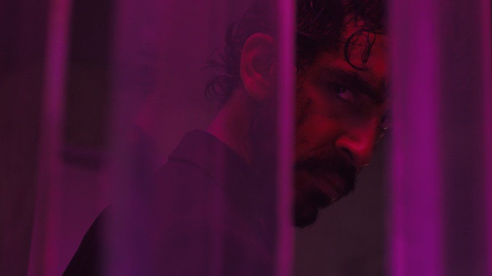 Dev Patel as 'Kid' in Monkey Man. Dev, a 33-year-old British Asian Man, looks at the camera with a sinister expression over his shoulder. A few locks of his curly dark hair flop over his face and he's wearing a dark blazer, facing away from the camera. He's lit by purple lighting and is slightly obscured by thin purple columns