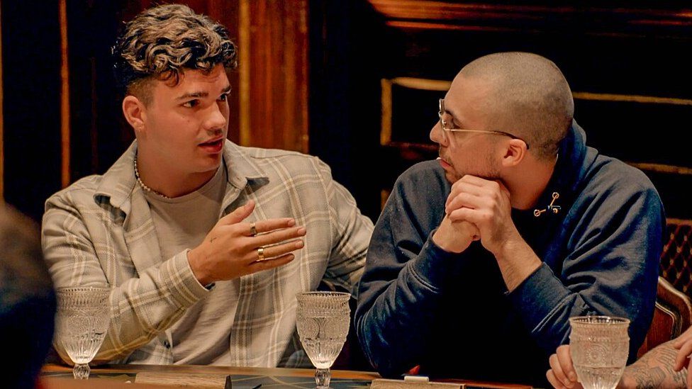 Harry and Zack at the Round Table in The Traitors. Harry is a 22-year-old white man with curly brown hair. He wears a grey checked shirt unbuttoned over a grey T-shirt. He has his left arm behind Zack while gesturing at him with his right. Zack is a 27-year-old white man with a shaved head. He wears glasses and a blue hoodie and looks at Harry, his hands under his chin. They are pictured inside the castle with dark wooden carved features and hanging lights. They each have a glass of water in front of them.