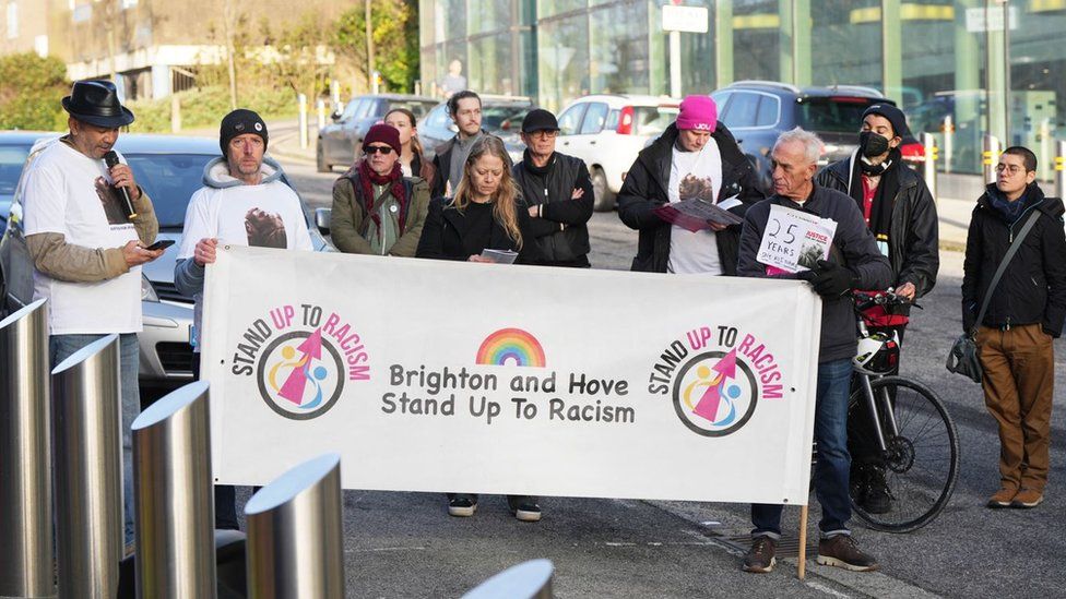 A group of people wearing t-shirts showing the face of Jay Abatan and carrying a banner which reads Brighton adn Hove stand up to racism