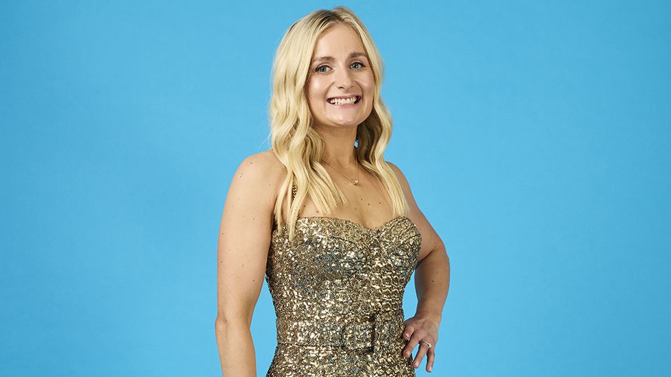 A woman in a golden, shoulderless, sparkly dress stands in front of a light blue background, smiling widely with one hand resting on her hip.
