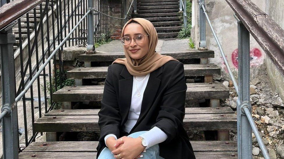 Female wearing beige headscarf, white top and a black blazer, sitting outside on wooden steps. There are wooden hand rails above metal hand rails. The background behind Amirah is of wooden steps with either side of concrete walls.