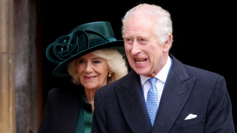 King Charles, accompanied by his wife Queen Camilla, smiles outside St George's Chapel in Windsor