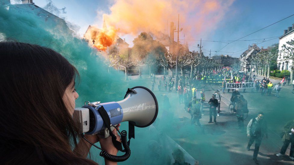 A protester speaks through a megaphone as smoke from coloured smoke bombs billows near people taking part in the annual May Day rally in Strasbourg