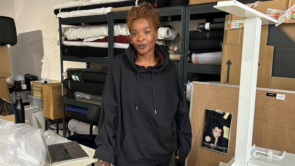 Torishéju Dumi pictured in her studio in Dalston, east London. Torishéju is a 31-year-old black woman with freckled skin and curly ginger hair which she wears tied back in a bun. She's wearing an oversized black hoodie and gold hoop earrings. Behind her, rolls of fabric are stored on shelves and there are boxes around her. She rests her right hand on a large table next to her which has an open laptop and some plastic wrapping on it