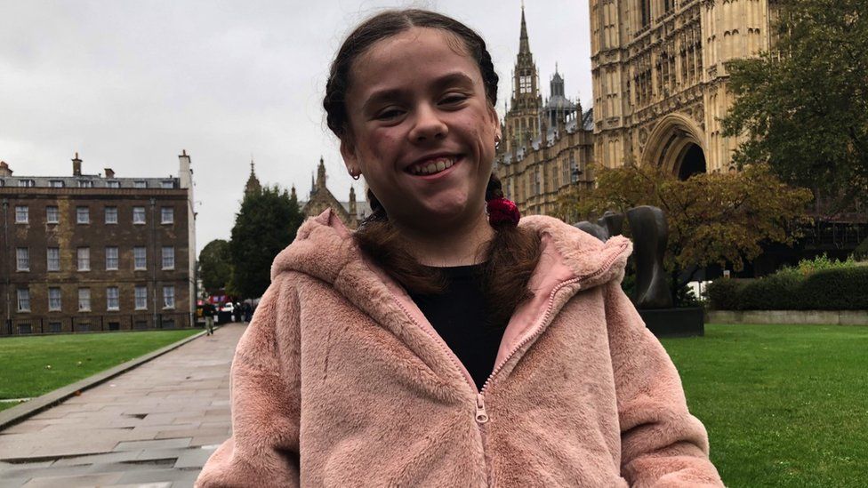 Carmela in Westminster before collecting her award. She is a 9 year old girl with brunette hair in two braids. She is wearing a pink fleece. She is looking directly at the camera with a big smile on her face. The Houses of Parliament can be seen in the distance behind her.
