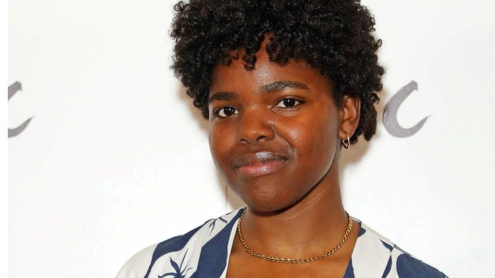 Francesca Amewudah-Rivers. Francesca is a black woman in her 20s with short afro hair and brown eyes. She smiles at the camera, wearing a blue and white blouse and small gold hooped earrings and gold chain necklace.