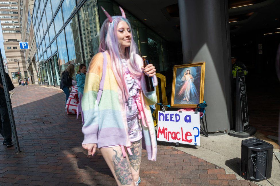 Outside the Marriott Copley hotel, a woman in a long pink and blue wig - wearing pink horns and a rainbow cardigan - smiles as she walks past a sign that reads: "Need a miracle?" and a picture of Jesus.