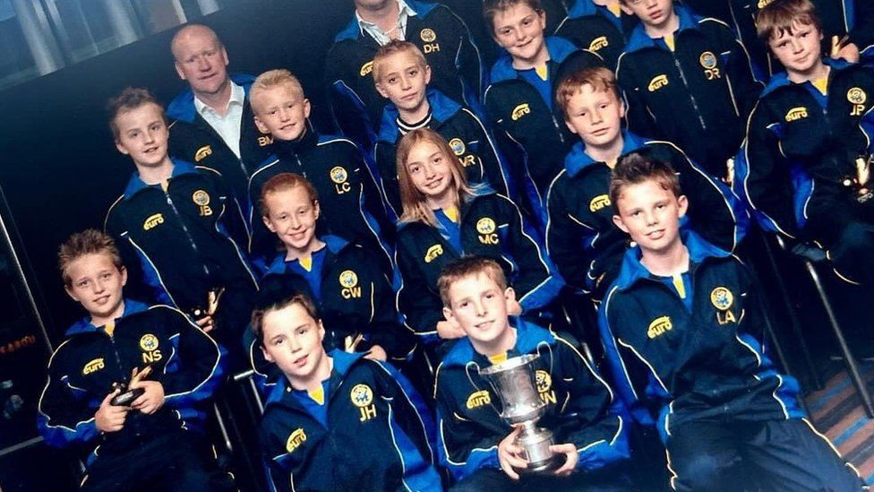 West Hallam juniors football team, a group of young children sit in blue tracksuits. One of the boys is holding a trophy. Maddy is the only girl in the photo.