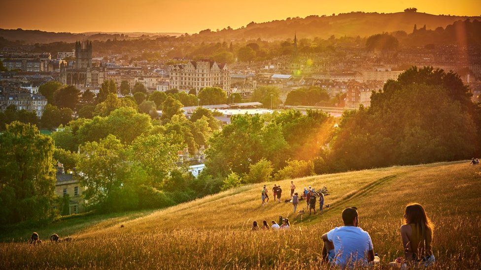 An orange evening sunset in Bath looking over hills. In the bottom right corner there is a couple sat together looking at the view, facing away from the camera