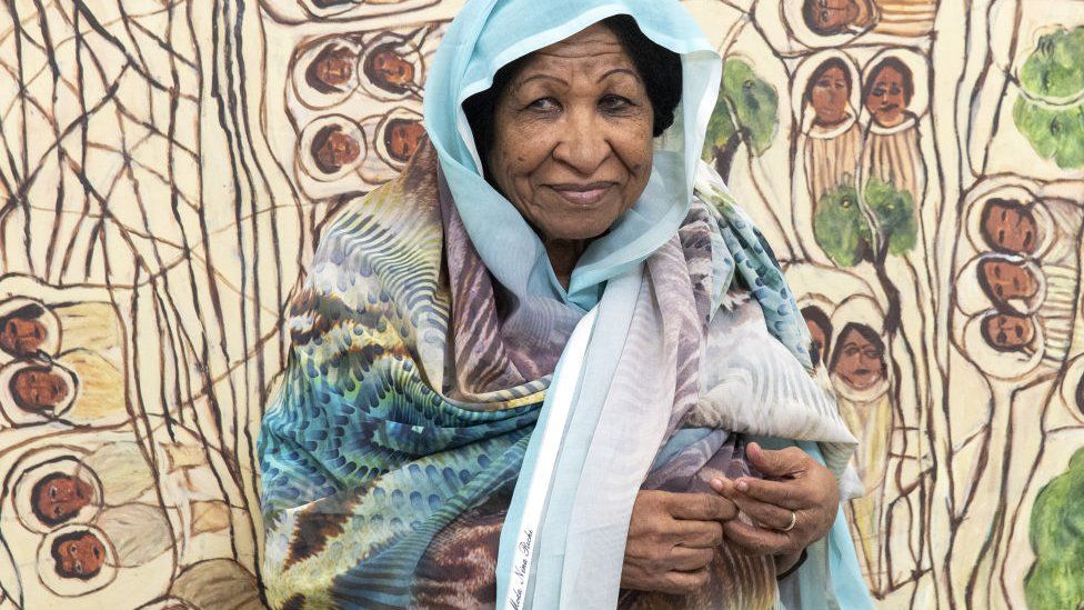 Kamala Ibrahim Ishag wearing a blue headscarf posing for a photo in front of artwork.