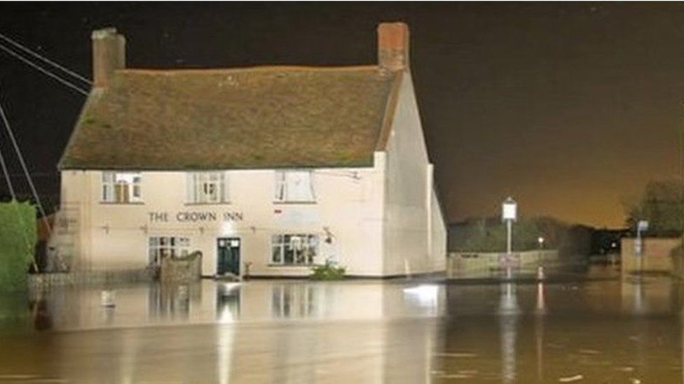 Two-storey pub "The Crown Inn" alongside a road which is covered in flood water.