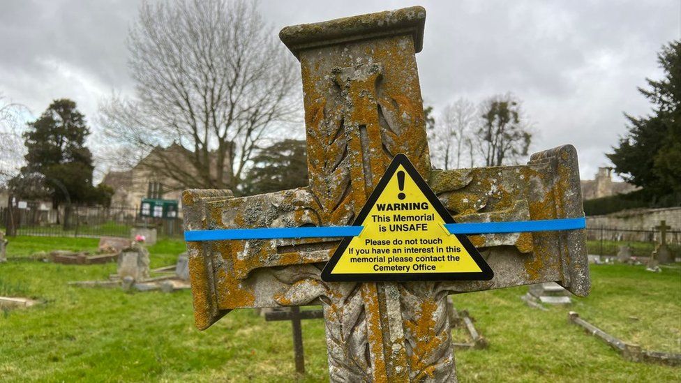 A cross headstone with a yellow warning taped onto it with blue tape. The warning reads: "Warning! This memorial is unsafe. Do not tamper with temporary support. Please contact Cemetery Office."