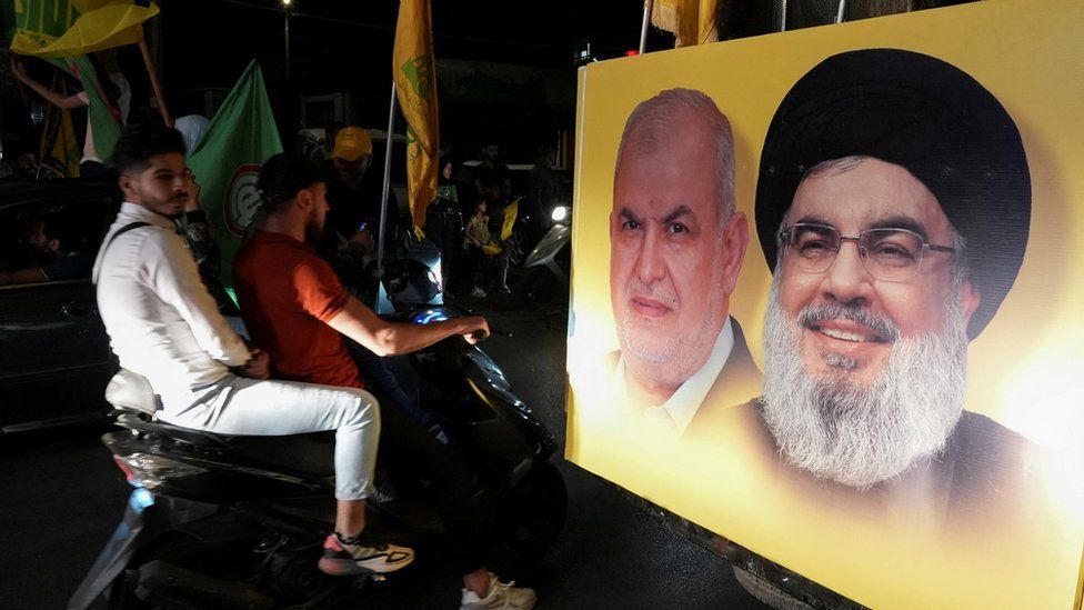Сторонники «Хизбаллы» несут флаги «Хизбаллы» и движения «Амаль», когда они едут в колонне мимо плаката с изображением лидера «Хезболлы» Хасана Насраллы и Мохамеда Раада, главы парламентского блока «Хезболлы», в Набатии, Ливан (15 мая 2022 г.)
