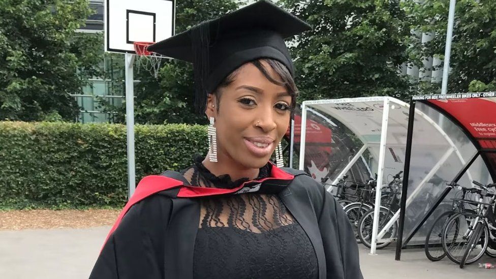 Rebecca Ikumelo, pictured at her graduation. Rebecca is a black woman in her 20s, she wears a mortar board and black university robes with a red trim over a black dress with lace details. She has short hair poking out beneath her hat and wars long dangly earrings and a nose ring. She is smiling at the camera and is pictured outside in front of a basketball hoop and bike shelter