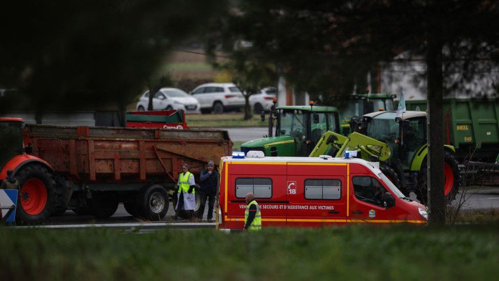 Rescuers stand by a firefighter vehicle, parked near the location where a farmer was killed and her husband and teenage daughter were seriously injured when they were run over at dawn on January 23, 2024