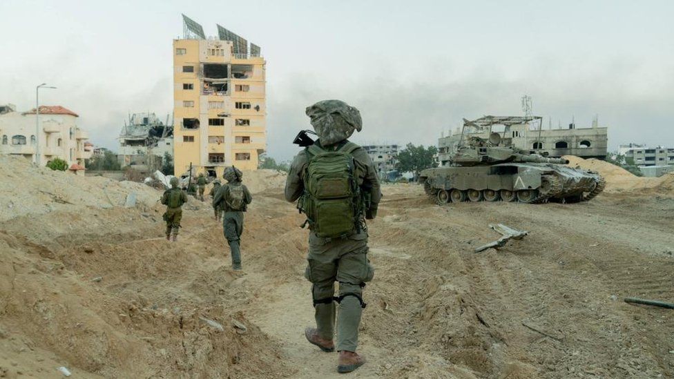 Israeli soldiers on patrol in the Gaza Strip. They are seen walking in single file across muddy ground towards a number of damaged buildings.