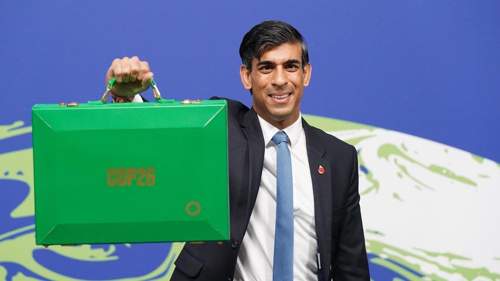 Риши Сунак держит чемодан с сокровищами, выкрашенный в зеленый цвет, перед мировым логотипом COP26