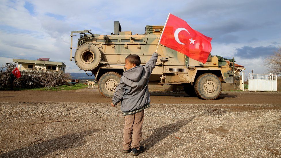 A photo taken from Turkey's Hatay province shows children greets soldiers with Turkish flags during Turkish military convoy consisting of approximately 300 armoured personnel carriers are on the way towards observation points in Syria's Idlib, on February 08, 2020 in Hatay, Turkey