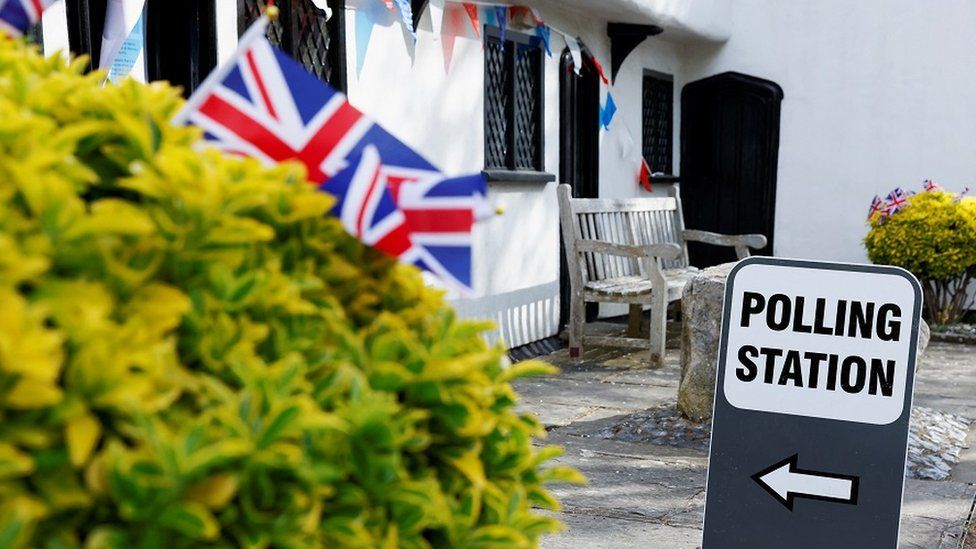 A polling station sign outside Barley Town House in Royston, Hertfordshire. In the foreground, some union jacks can be seen sticking out of a hedge