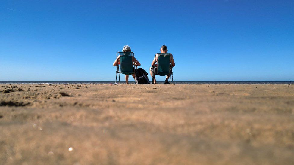 A man and a woman sit on deck chairs under blue skies on Blackpool beach