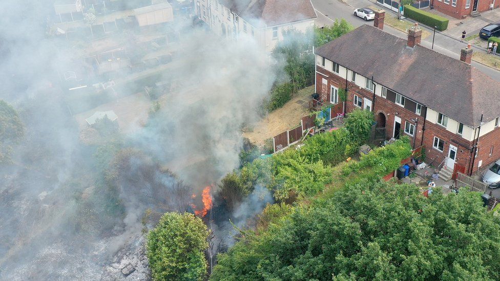 На этом виде с воздуха пожарные борются с лесным пожаром, охватившим близлежащие дома в районе Ширгрин в Шеффилде 20 июля 2022 года в Шеффилде, Англия.