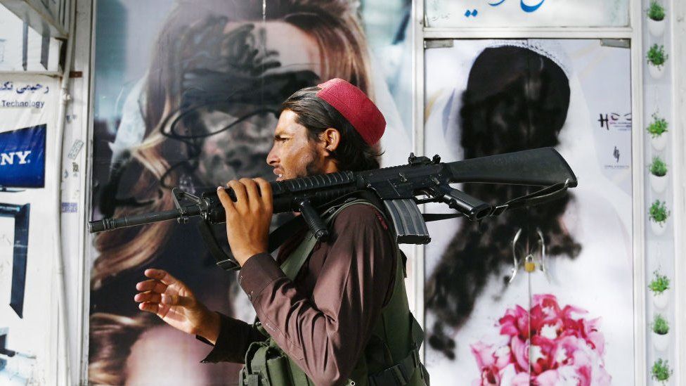 A Taliban fighter walks past a beauty salon carrying an M16 weapon.