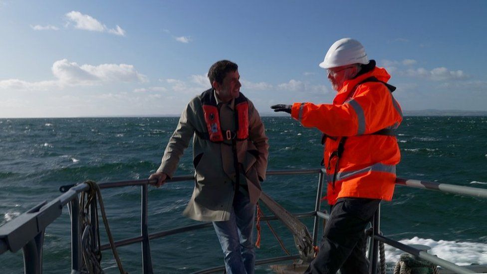 Climate editor Justin Rowlatt on a boat speaking with a man wearing orange high-vis and a white hard hat
